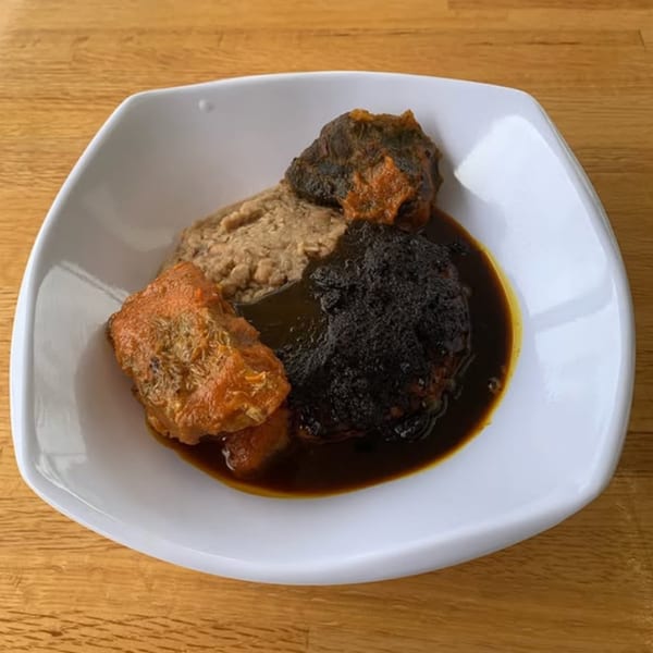 A white square bowl filled with T's Buka African stew, beans, and meat on a wooden table.