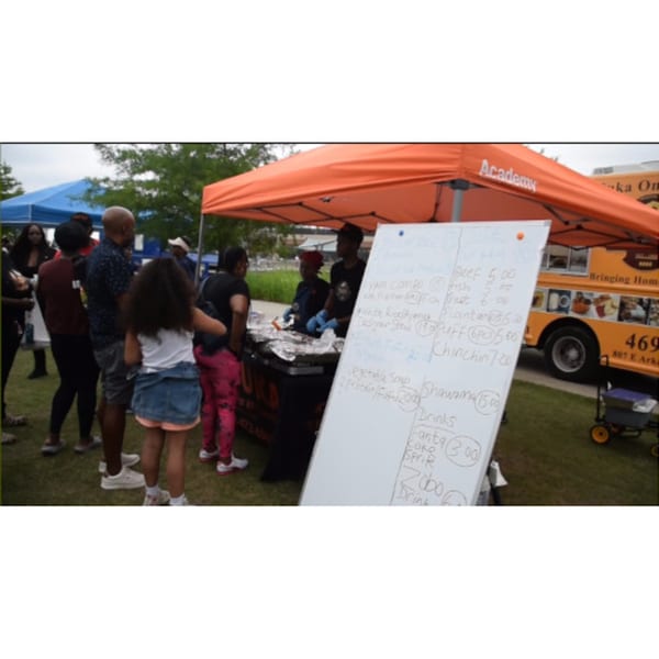 People gather around T's Buka food truck and tent with a menu board displaying African dishes.