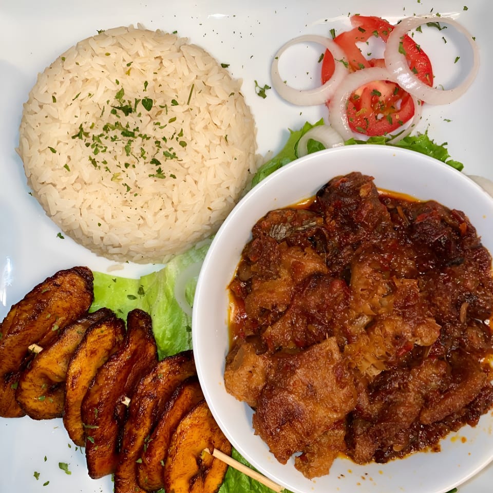 A plate of African food with white rice, stewed meat, fried plantains, and a side salad.